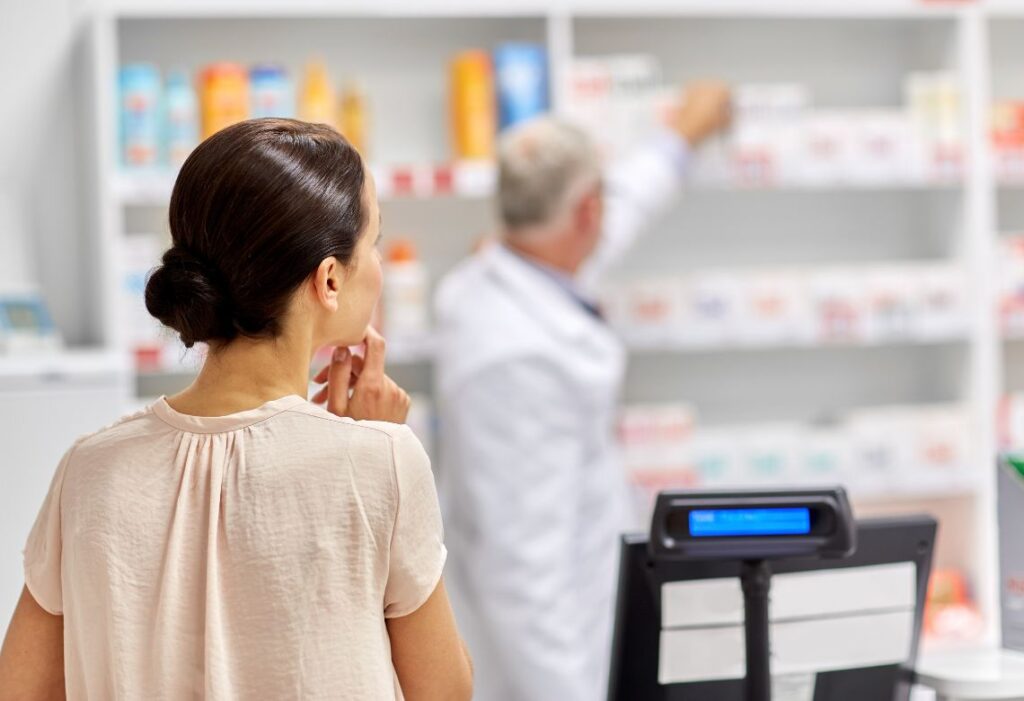 Mulher escolhendo remédio e farmacêutico ao fundo com a prateleira de remédios