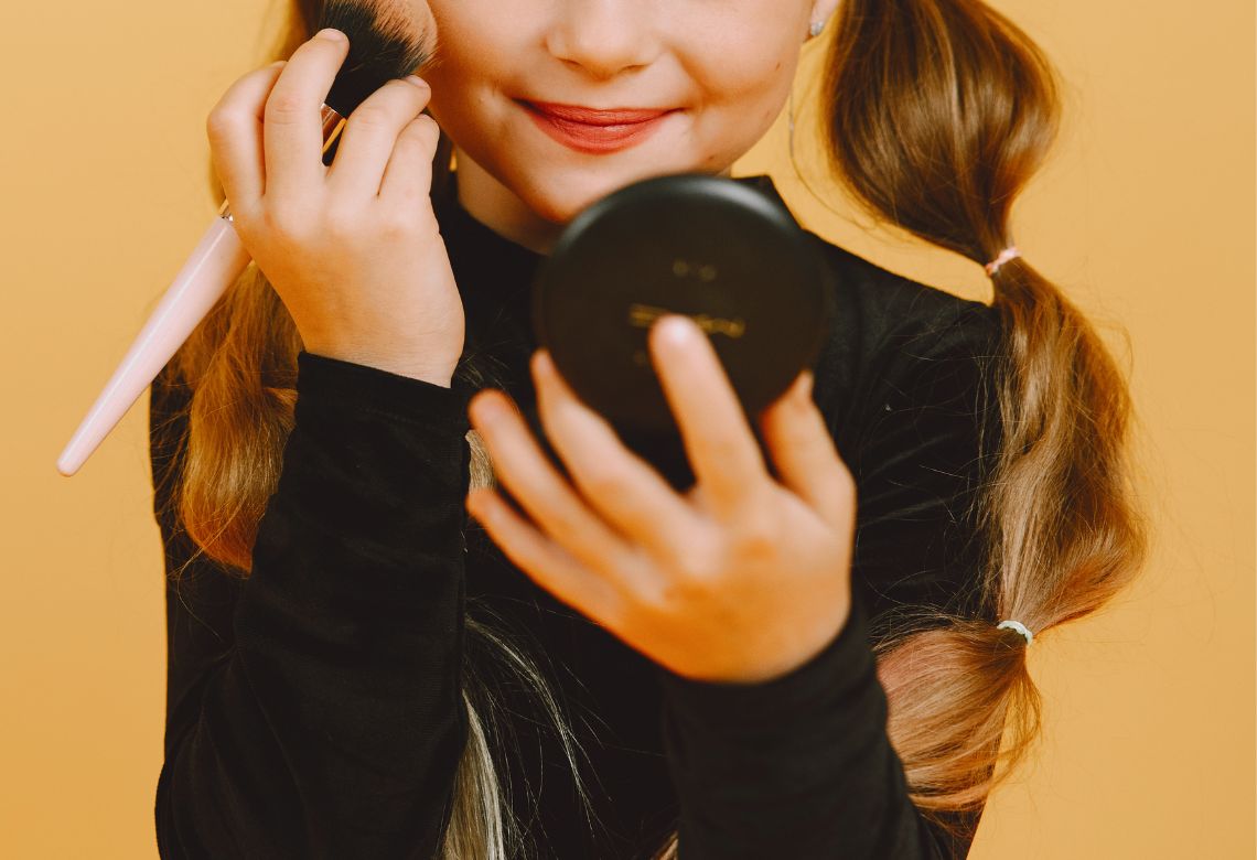 Menina se maquiando, segurando um blush com a mão esquerda, passando o blush nas bochechas e segurando um espelho com a mão direita. Ela está vestida de preto e a parede ao fundo é laranja pastel.